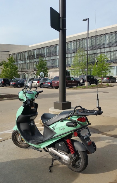 Photograph of a scooter in the parking lot of an office building. The scooter is closest to the viewer and is the largest item in the field of view, with a number of cars in the midrange view, and an office building in the distance.