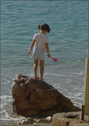 Photograph of a young girl holding a little plastic shovel, standing on a large rock at the edge of the water at the beach.